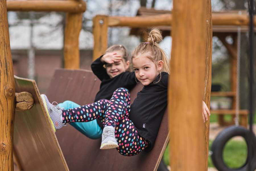 Two little girls on the playground are suspended in a wooden climbing frame, wearing black t-shirts and colorful leggings